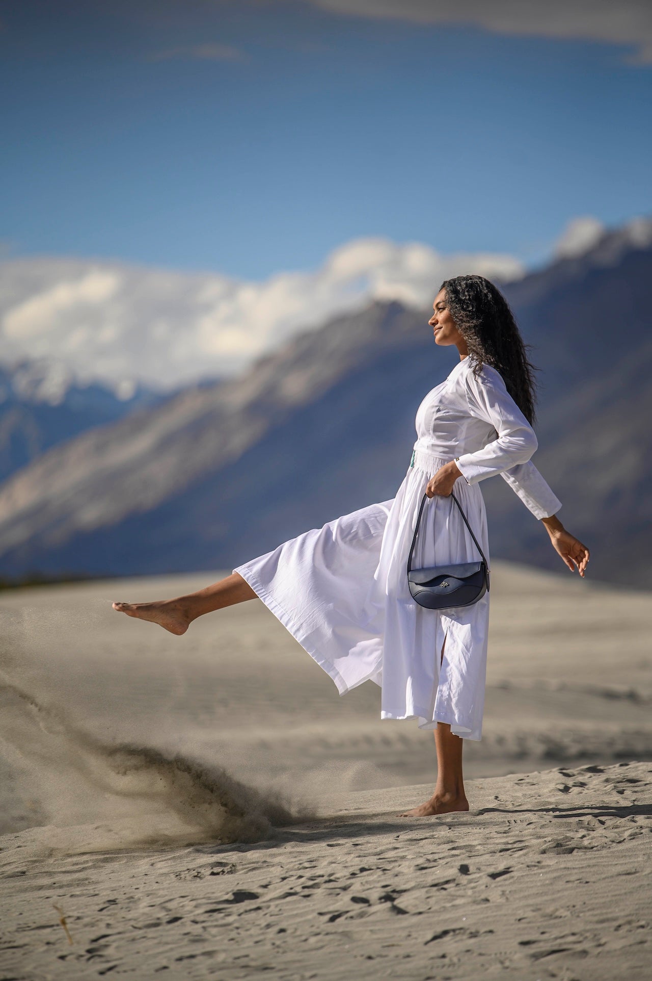 Woman in a white dress standing on sand with mountains in the background