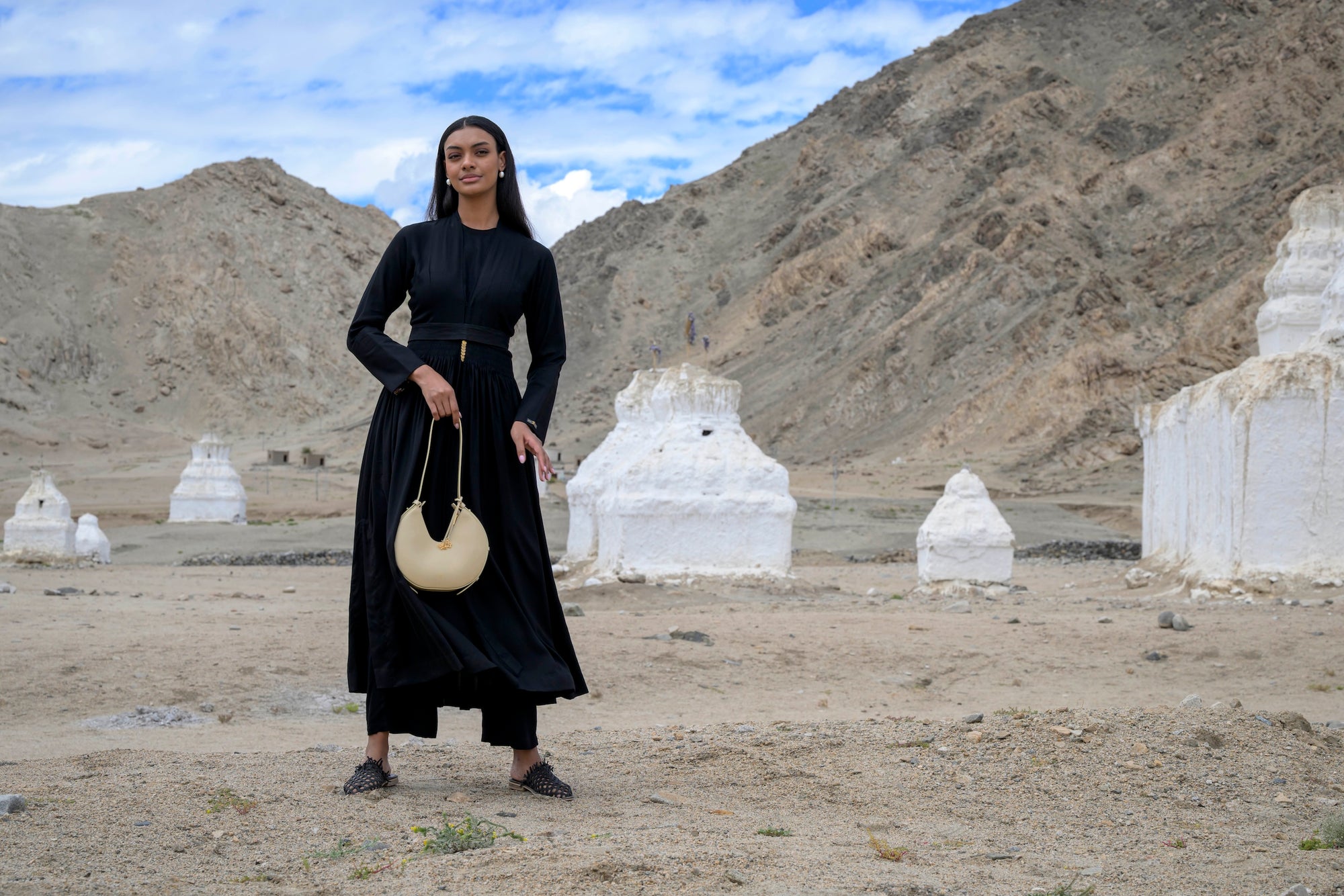 Woman in a black dress holding a beige linearity bag standing in a desert landscape with white structures.