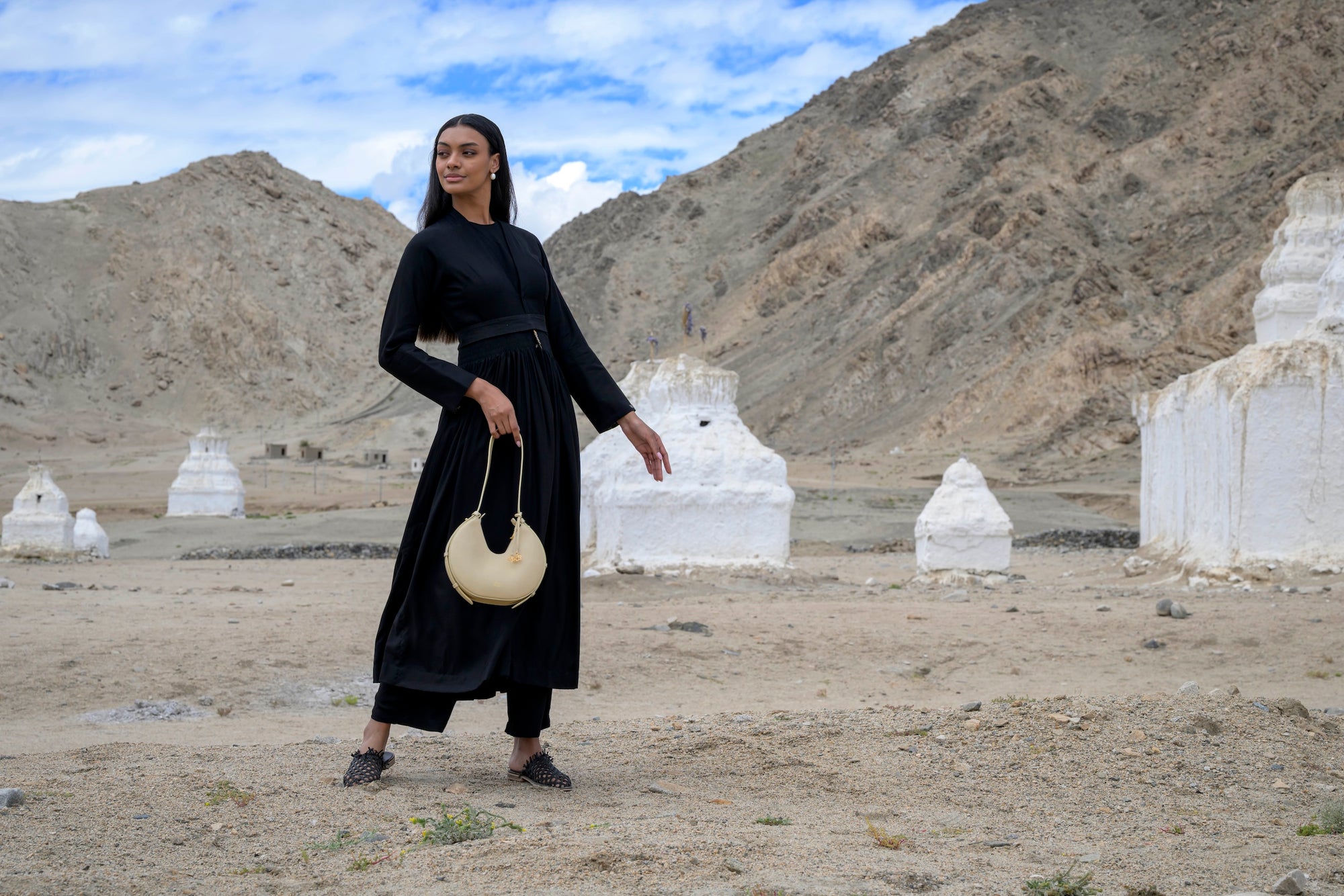 Woman in a black outfit holding a beige linearity bag in a desert-like landscape with white structures and mountains.