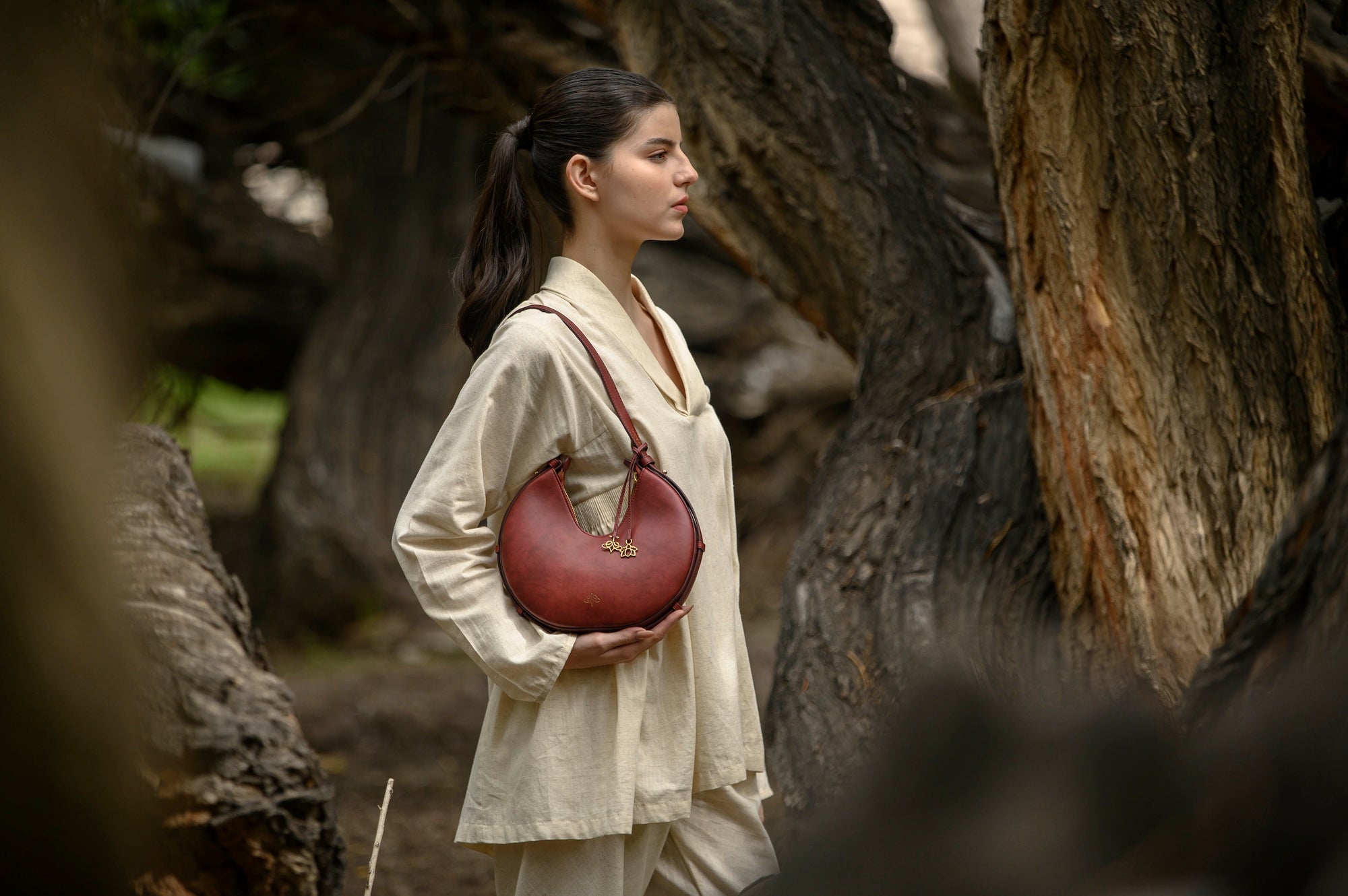 Woman holding a bordeaux linearity handbag in a natural setting with trees.