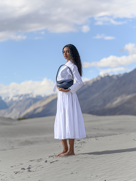 Model in a white dress holding the Linearity dark blue bag on a sandy landscape with mountains in the background