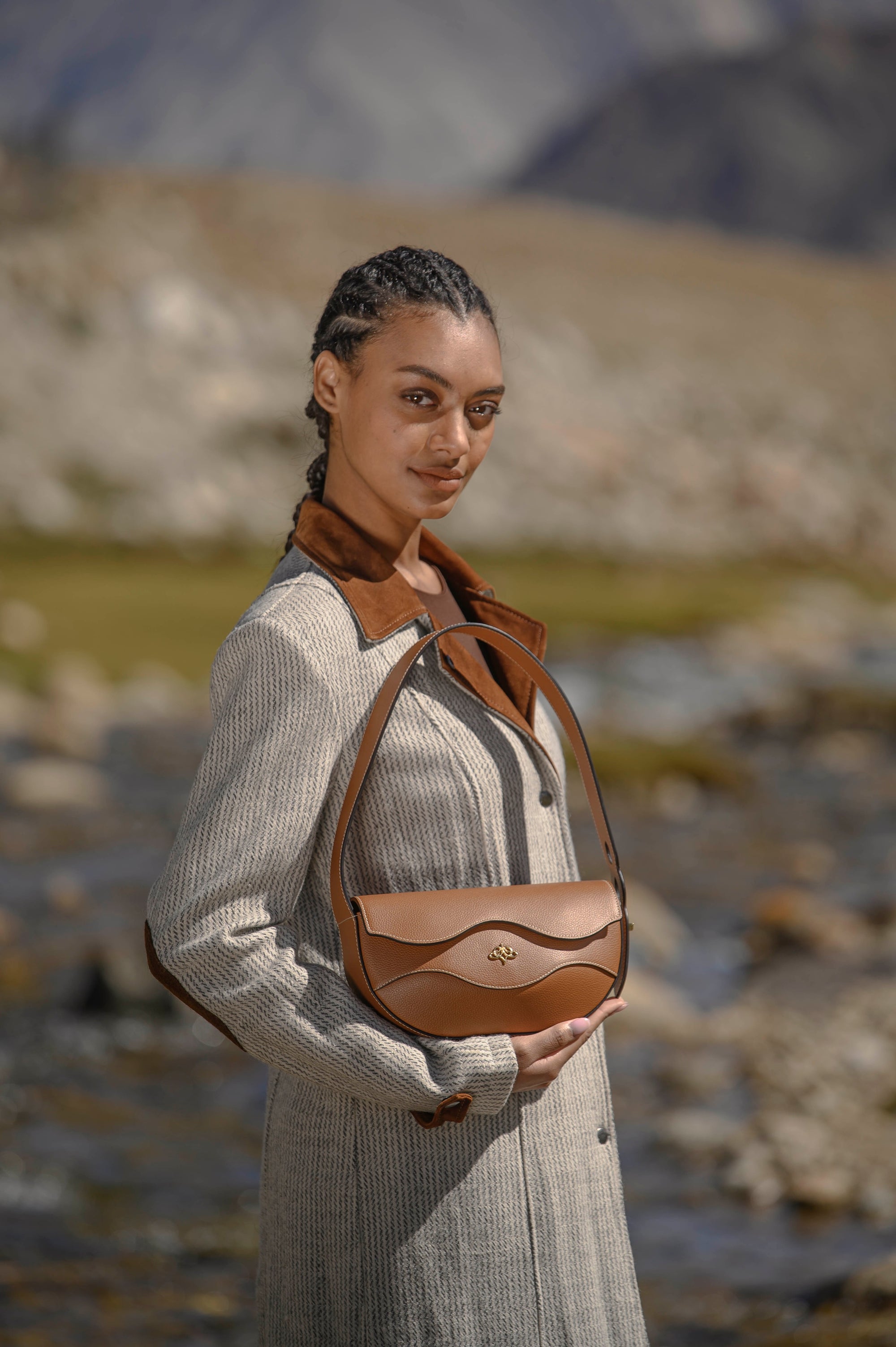 Woman holding a brown Linearity handbag in a natural setting with mountains in the background