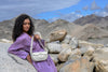 Woman in a purple dress holding an Ascot grey linearity bag on rocky terrain with mountains in the background