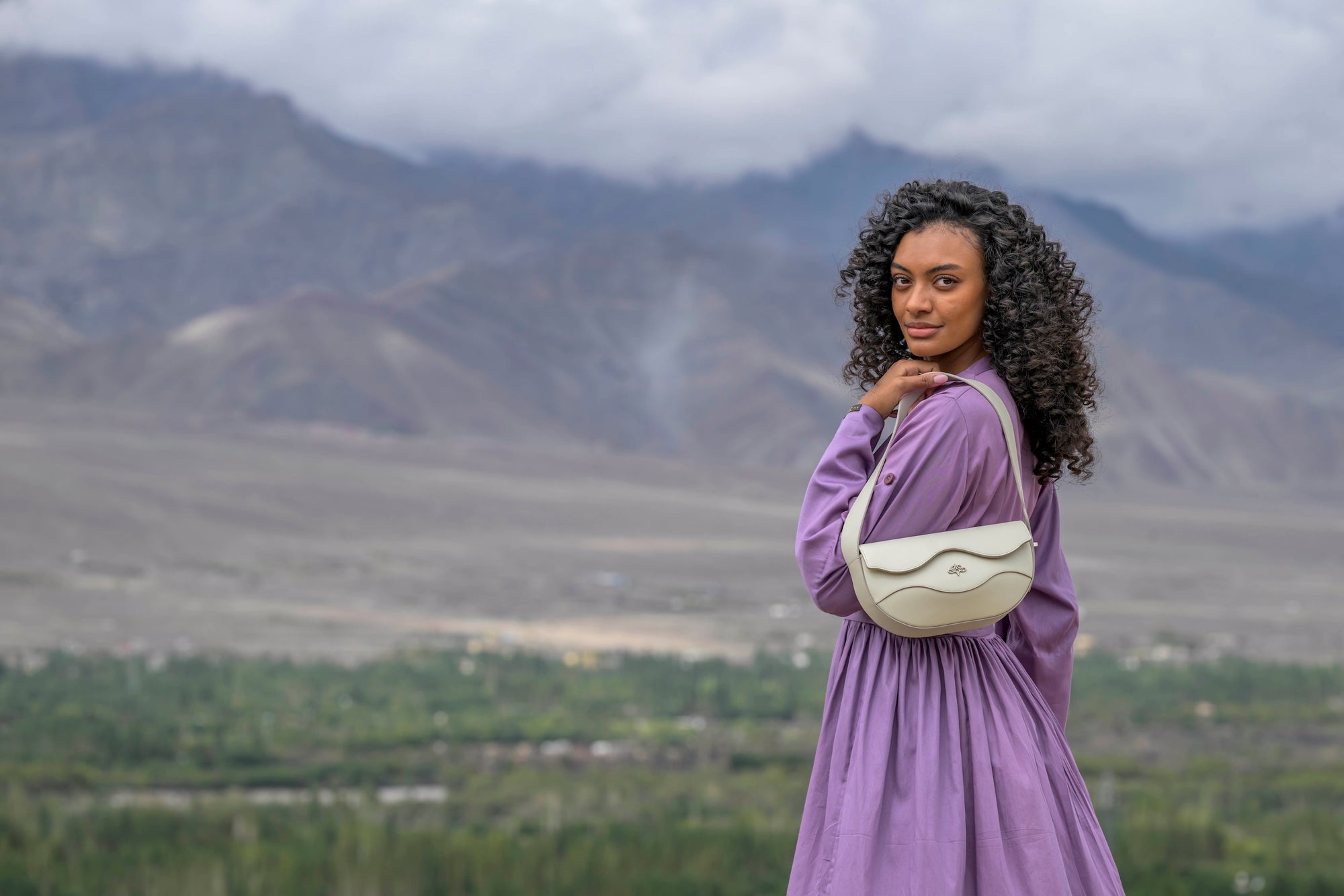 Woman in a purple dress with a white linearity bag standing in a scenic landscape with mountains and greenery.