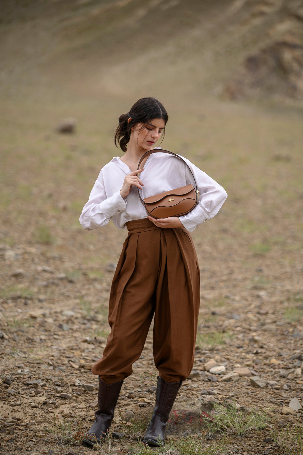 Woman in a white blouse and brown pants holding a brown linearity bag in a desert-like setting