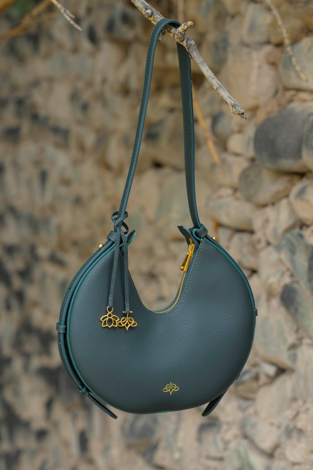 Dark green Linearity handbag with gold accents hanging on a branch against a stone wall.